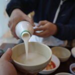 A person pouring traditional Korean makgeolli into a bowl at a restaurant, with a spread of Korean side dishes on the table