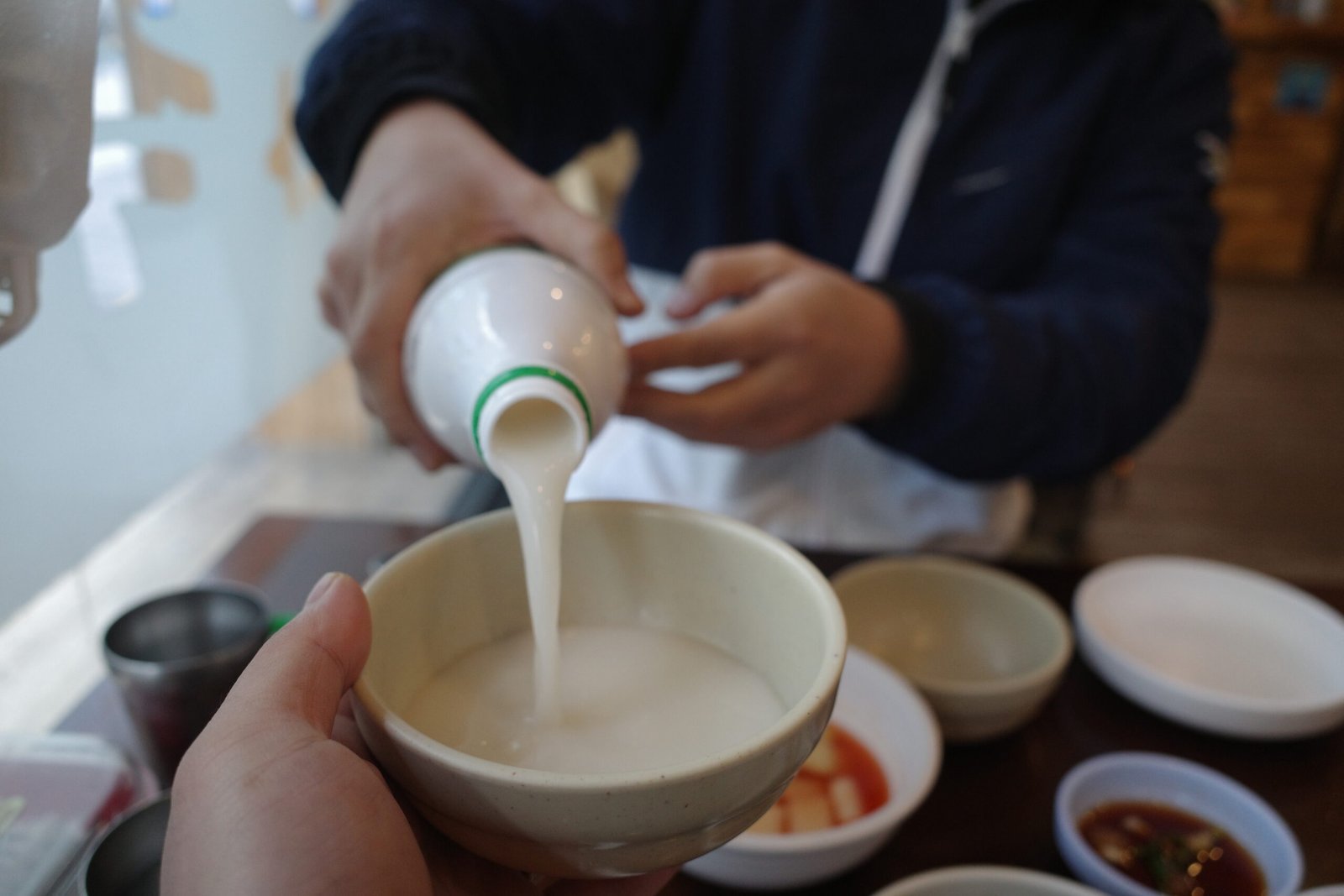 A person pouring traditional Korean makgeolli into a bowl at a restaurant, with a spread of Korean side dishes on the table